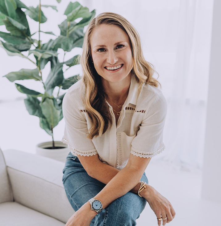 Smiling woman sitting on a light-colored couch in a bright room with a large green plant in the background.