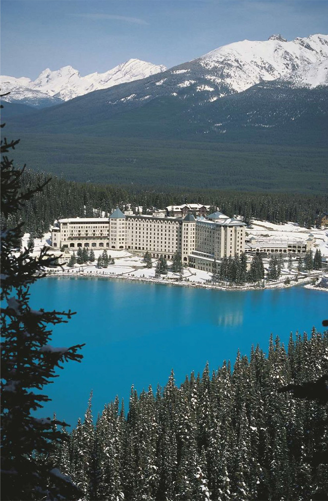 Large hotel complex by a bright blue lake with snow-covered mountains in the distance.