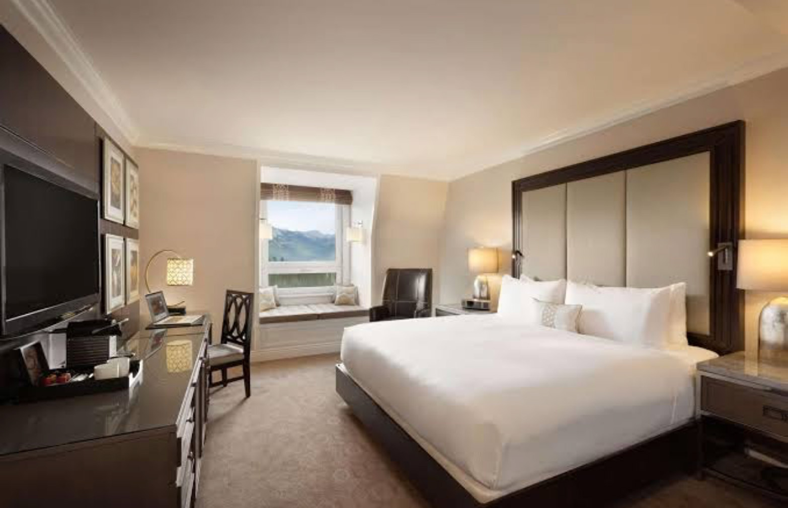 Spacious hotel room with a king-size bed, padded headboard, two nightstands, and a window seat overlooking mountains.