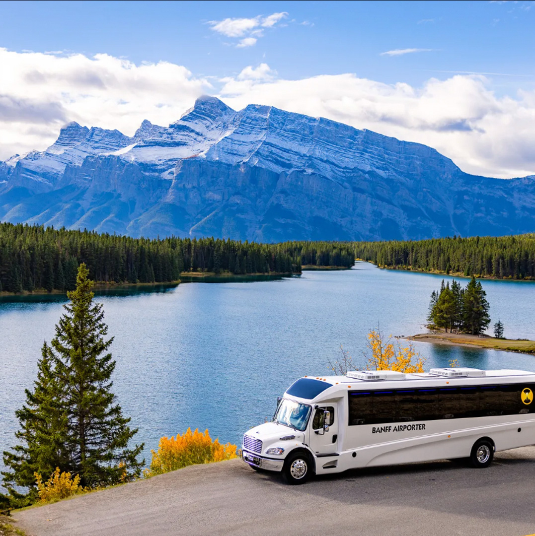 White Banff Airporter bus parked on a road by a blue lake, with evergreen trees and snow-capped mountains in the distance.