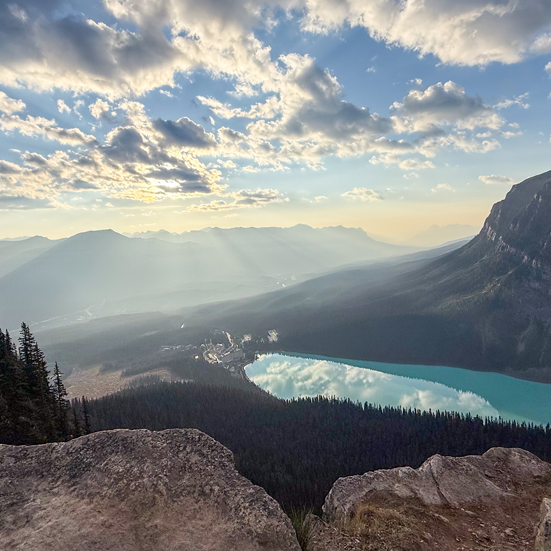 Mountain valley with a turquoise lake, dark forests, and rocky foreground under a partly cloudy sky at sunset/early morning light, distant ridges glowing.