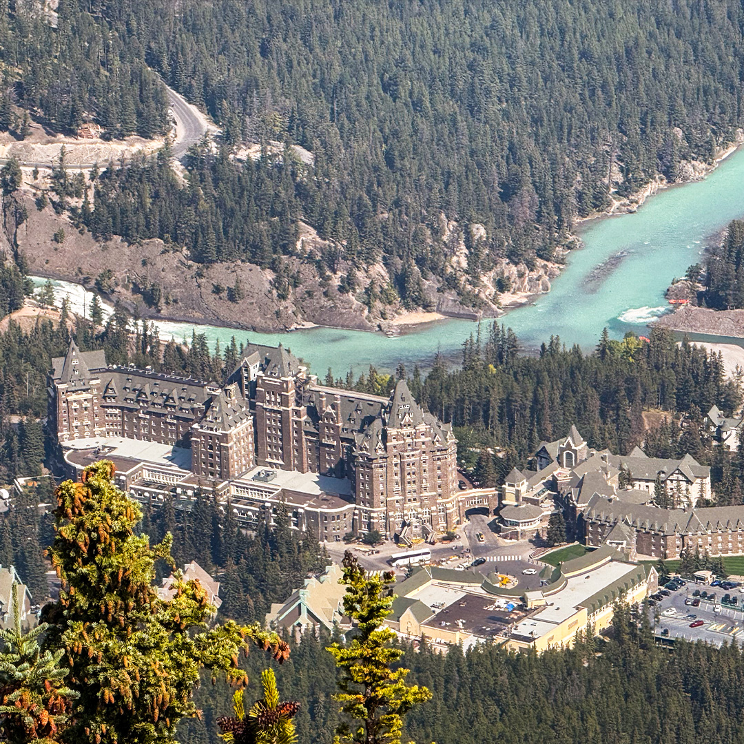 Castle-style resort nestled among evergreen trees beside a turquoise river in the mountains.