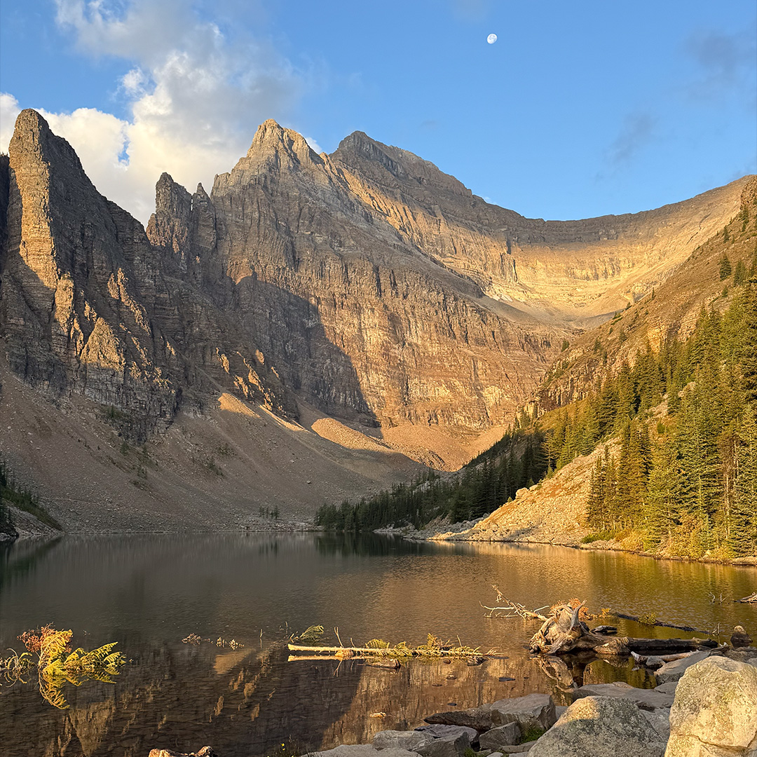 A tranquil alpine lake reflecting jagged mountain peaks under a clear blue sky with a small moon visible above. On the right, evergreen trees line the shore and rocky foregrounds frame the scene.