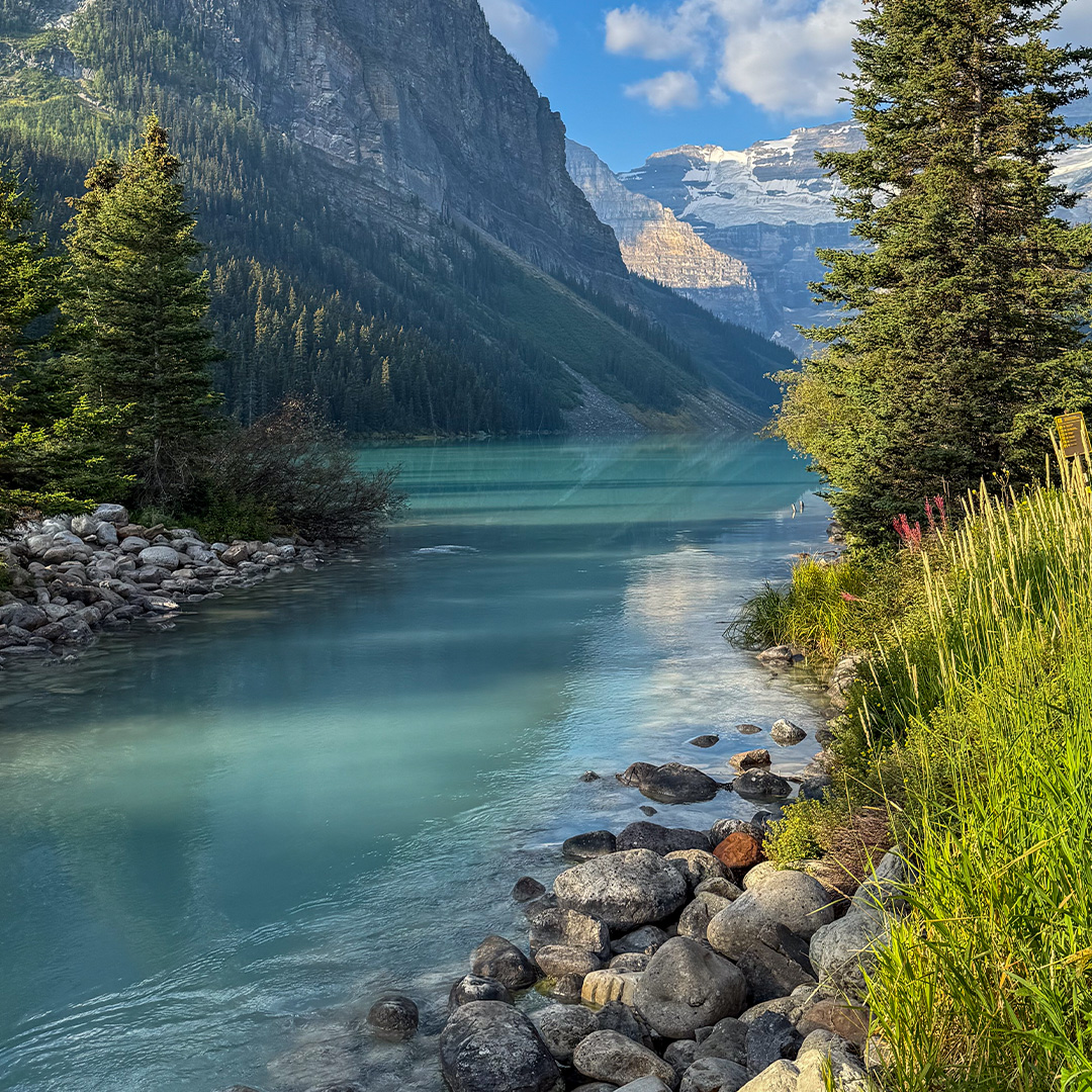 Turquoise alpine lake with a rocky shore, evergreen trees, and tall mountains under a clear blue sky.