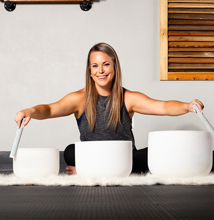 Smiling woman sitting on a rug, playing three white singing bowls with mallets in a meditation/yoga setting.