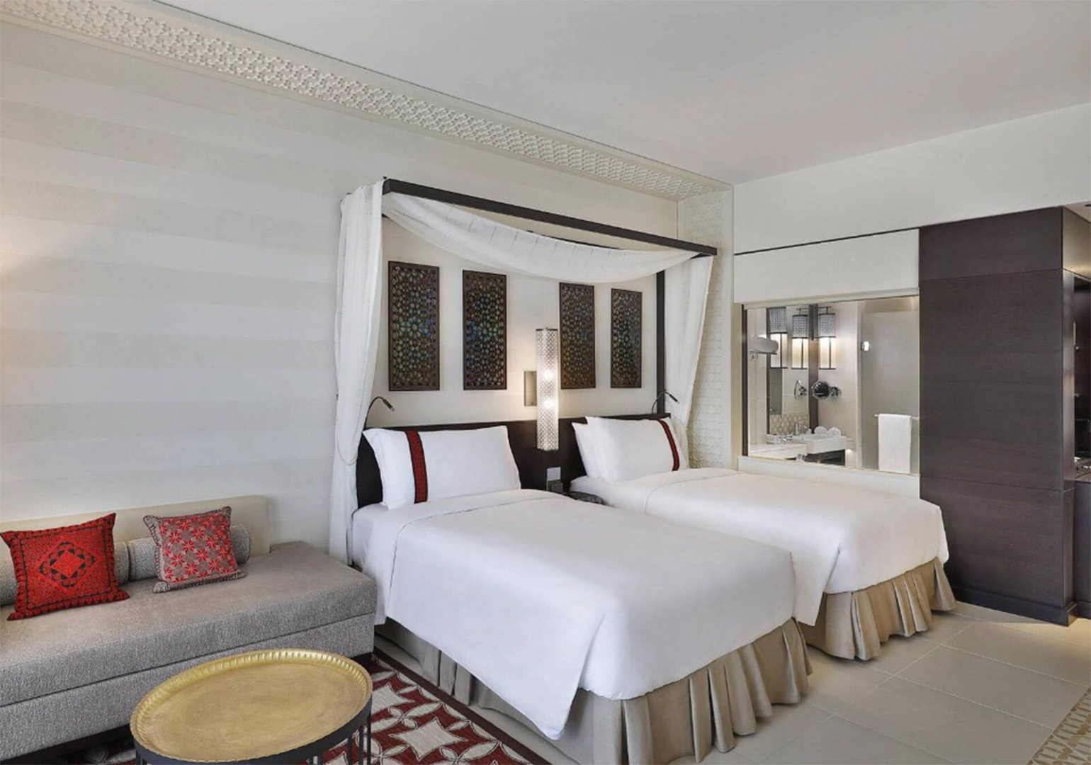 Twin-bed hotel room with a white canopy over the headboard, decorative wall panels, and a seating sofa with red pillows on the left edge.