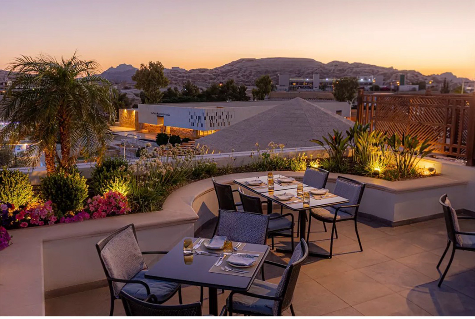 Rooftop restaurant patio at sunset with multiple dining tables set for guests and colorful plants along the edges, city hills in the distance.