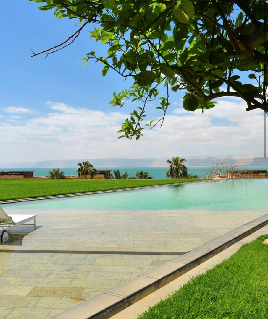 Poolside resort scene with turquoise water, green lawn, and distant palm trees under a blue sky