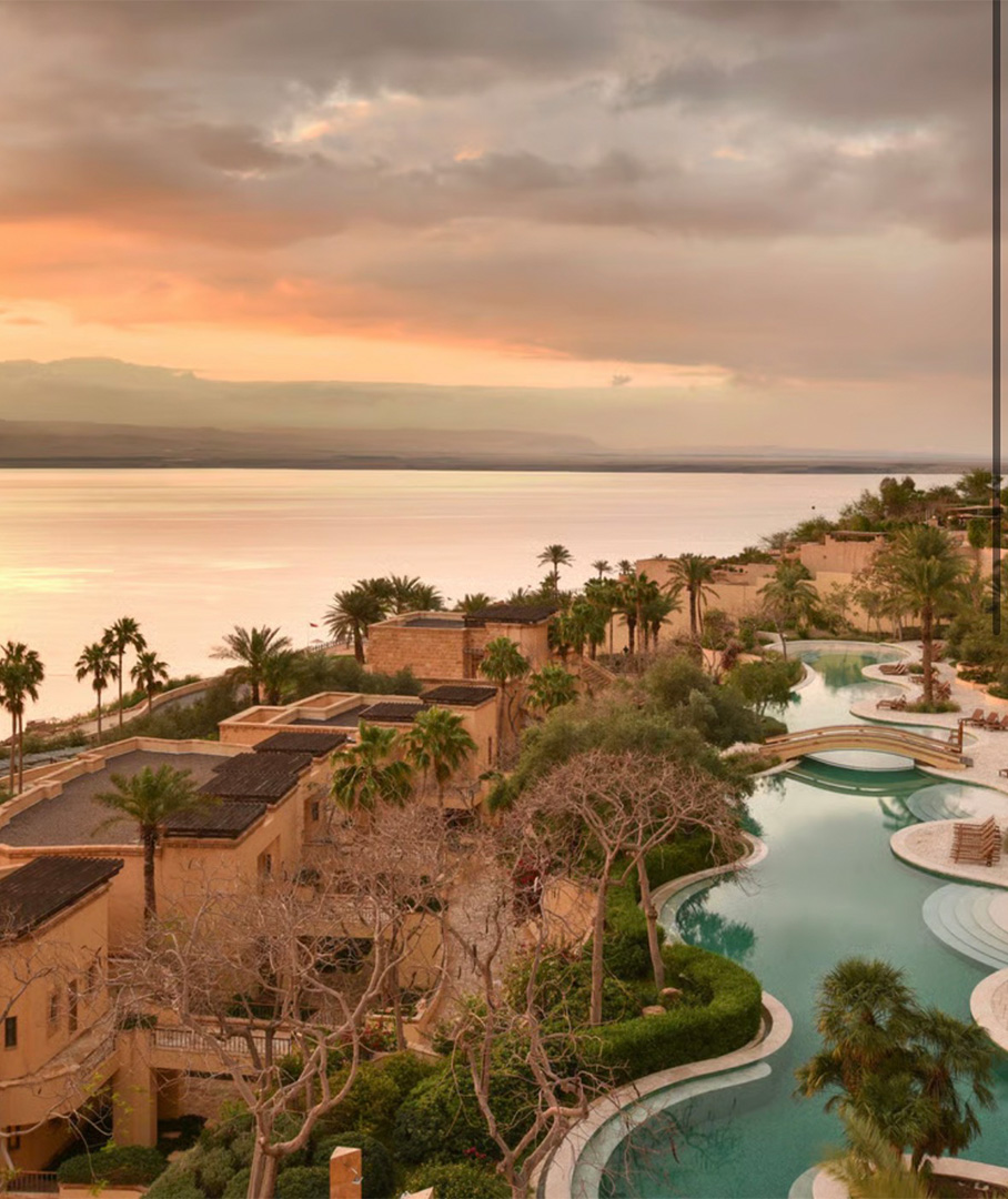 Sunset over a terracotta resort with palm trees and a winding pool along the shoreline.