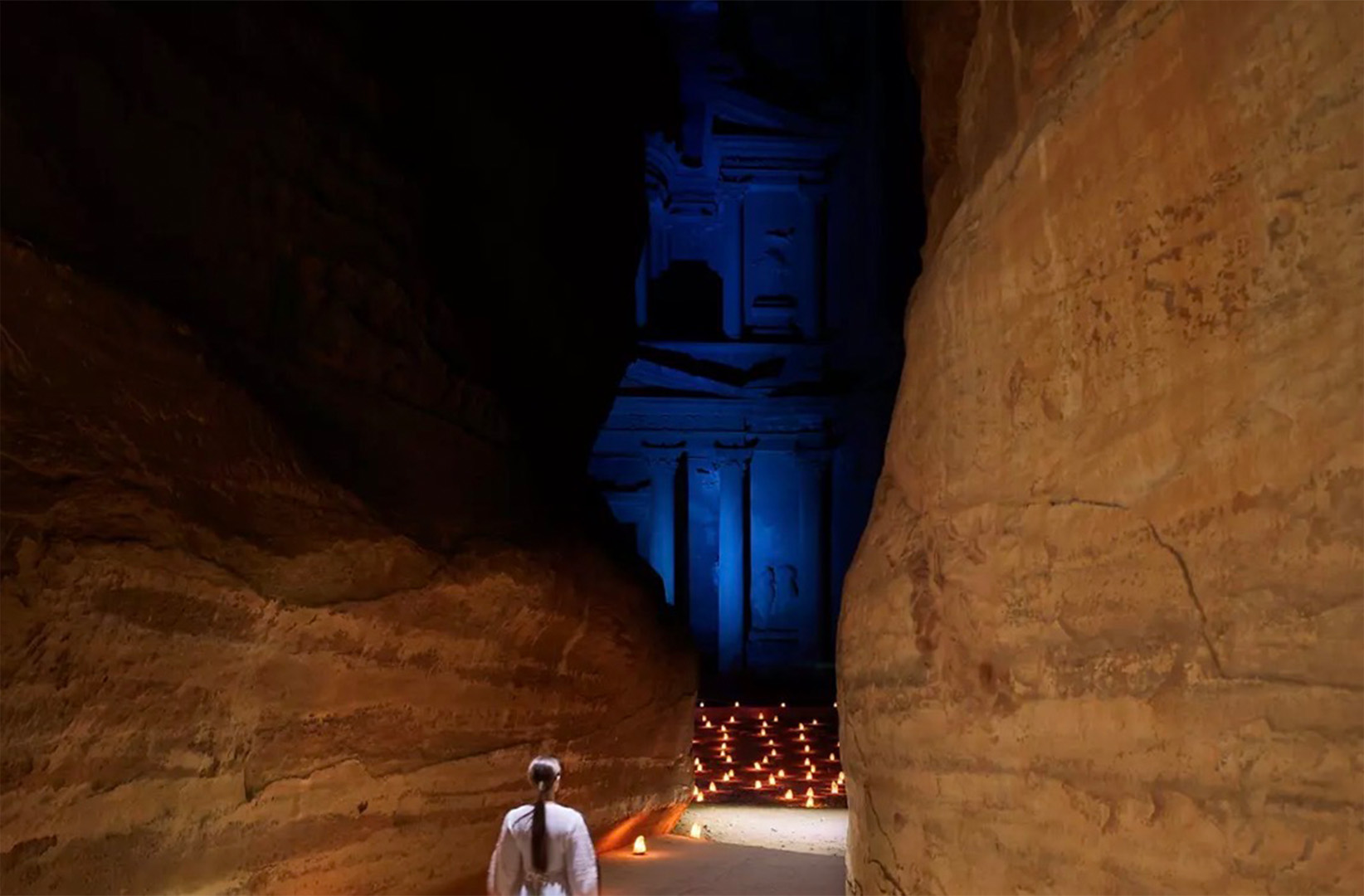 Inside a narrow cave, orange rock walls frame a blue-lit carved facade at the far end; candles line the floor, with a person in white nearby.