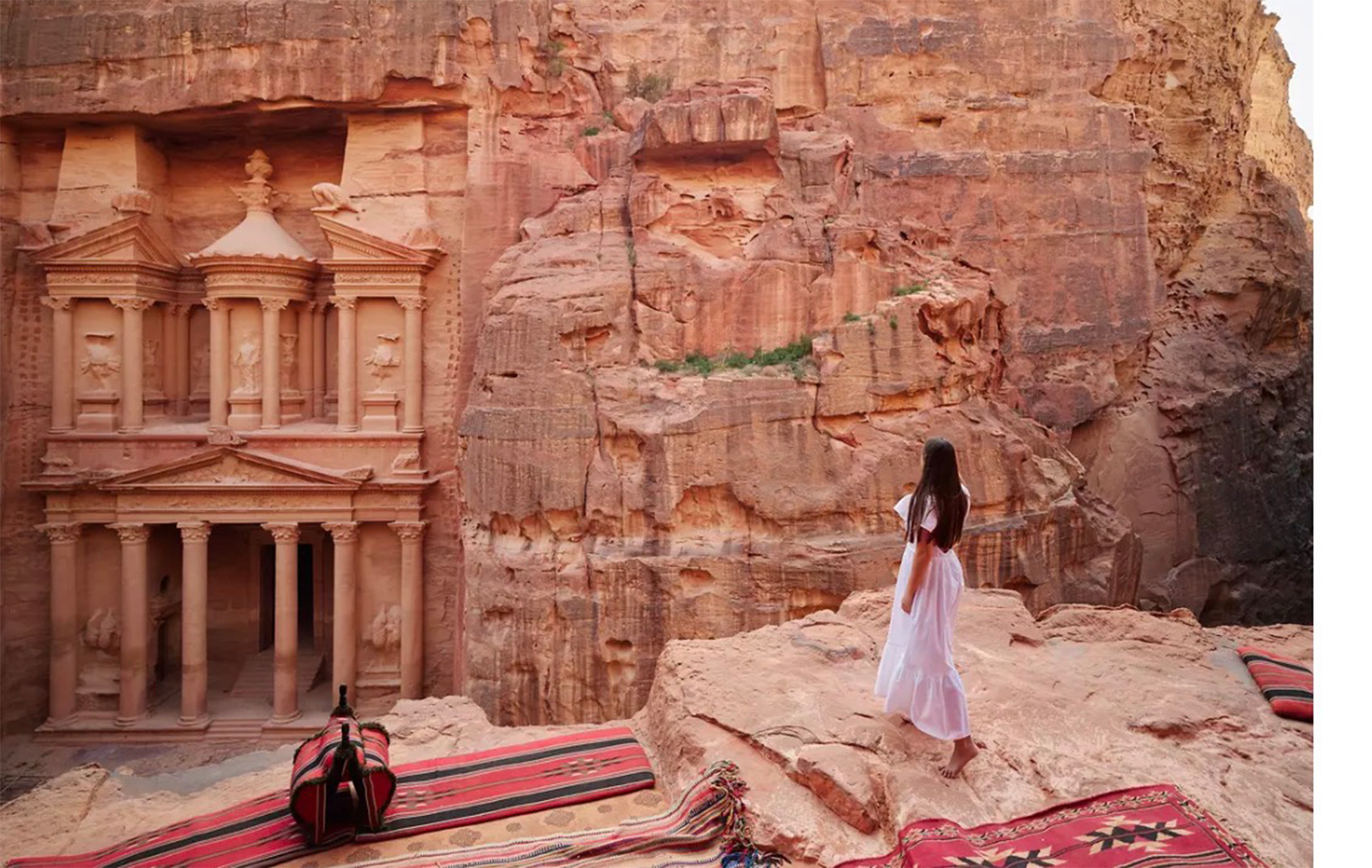 A woman in a white dress stands on a rocky ledge beside the Petra-like rock-cut temple, with colorful woven carpets spread on the ground.