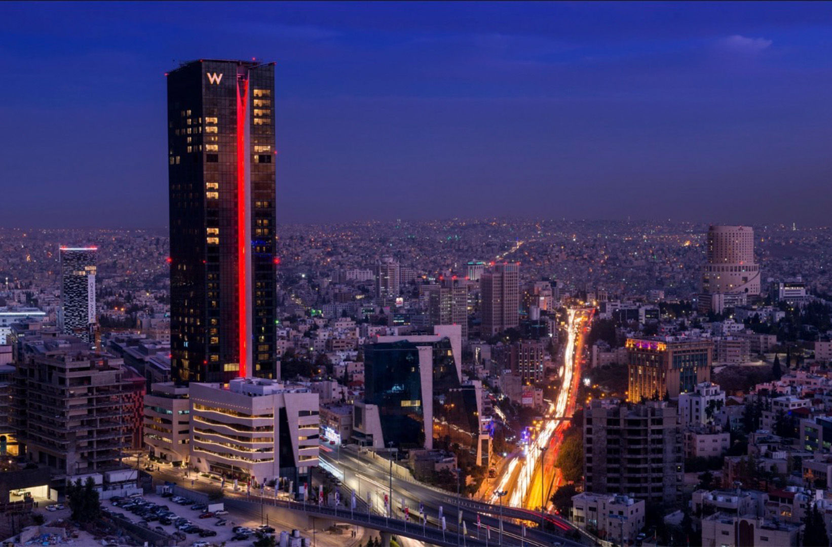 City skyline at dusk with a tall dark skyscraper lit by a red vertical stripe and a busy, illuminated highway below.