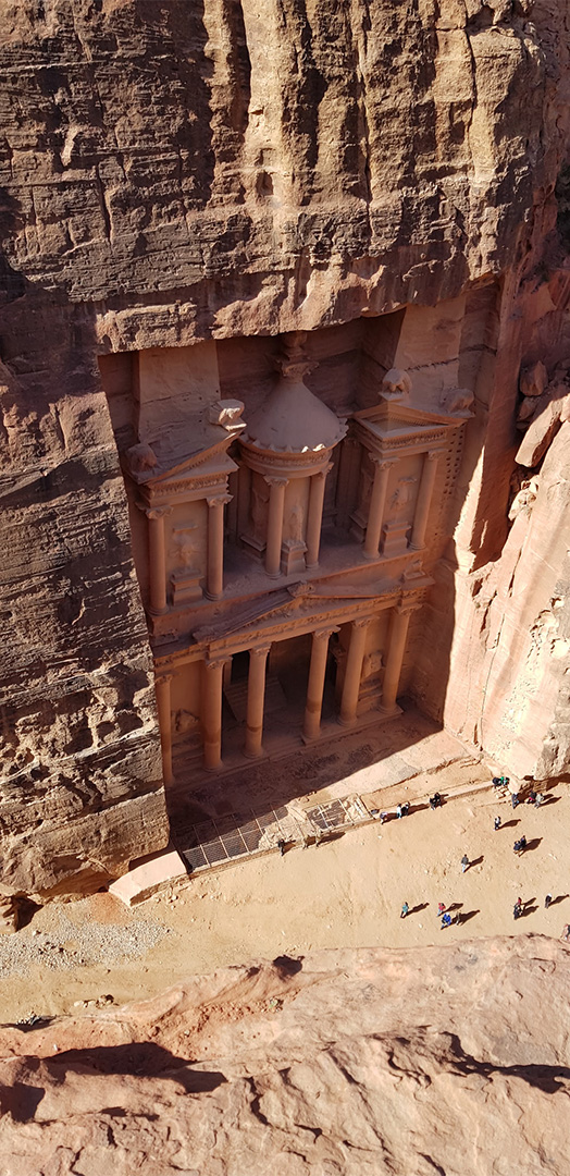 A sandstone cliff with a carved rock‑cut facade featuring three temple-like structures and columns, viewed from above with visitors at the base for scale.