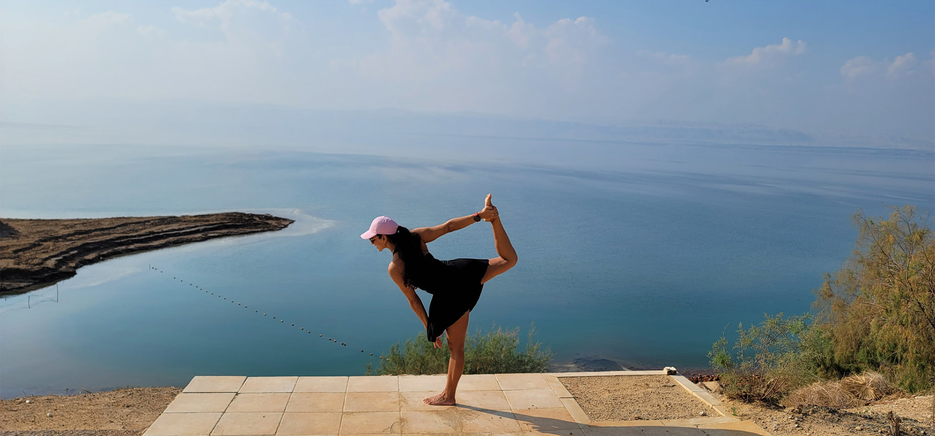 Woman in a pink cap and black outfit balances in a dancer pose on a tiled platform by a calm blue lake with distant hills.