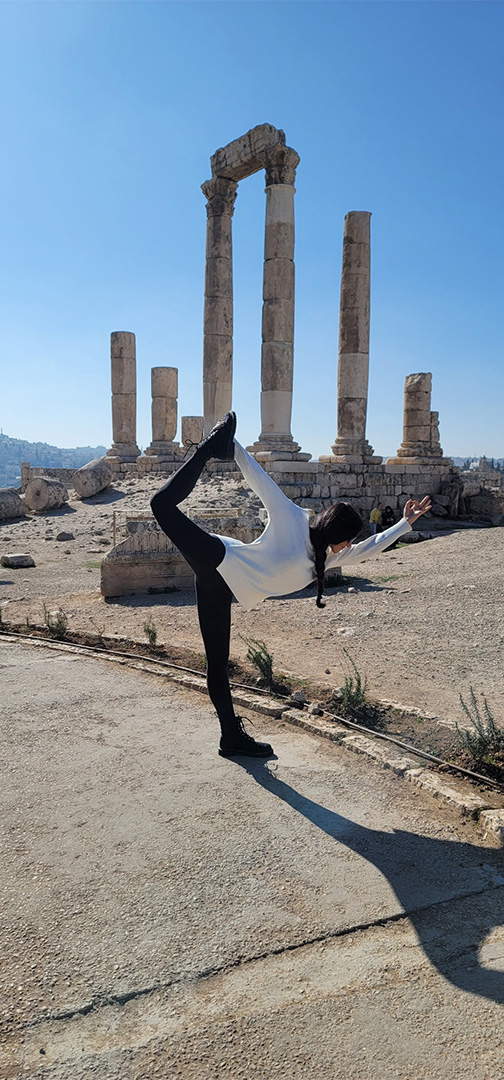 Person performing a yoga balance pose in front of ancient stone columns at a sunny archaeological site.